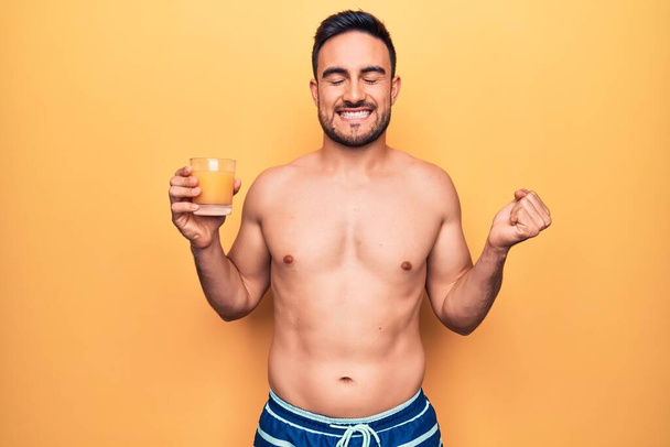 Junger gutaussehender Mann mit Bart im Urlaub in Badebekleidung, der ein Glas Orangensaft trinkt und stolz schreit, Sieg und Erfolg mit erhobenem Arm feiert - Foto, Bild
