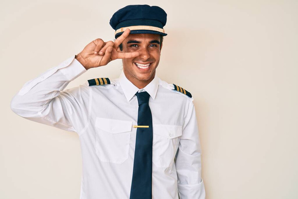 Junger hispanischer Mann in Flugzeugpilotenuniform macht Friedenssymbol mit den Fingern über dem Gesicht, lächelt fröhlich und zeigt den Sieg  - Foto, Bild