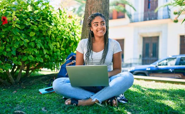 Genç Afrikalı Amerikalı öğrenci mutlu bir şekilde gülümsüyor. Üniversite kampüsünde çimlerin üzerinde bilgisayarla oturuyor. - Fotoğraf, Görsel
