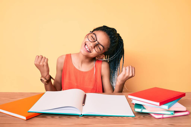 Young african american girl child with braids studying for school exam very happy and excited doing winner gesture with arms raised, smiling and screaming for success. celebration concept.  - Foto, imagen