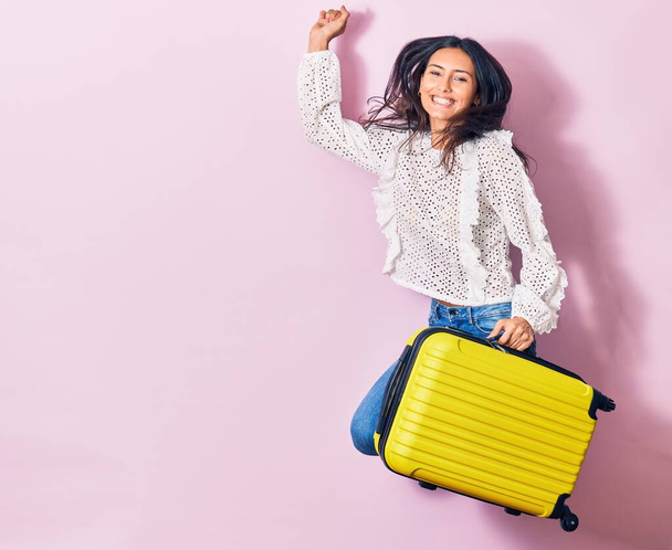 Young beautiful woman on vacation wearing summer clothes smiling happy. Jumping with smile on face holding cabin bag over isolated background - Fotó, kép