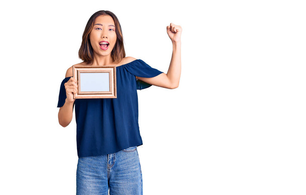 Young beautiful chinese girl holding empty frame screaming proud, celebrating victory and success very excited with raised arms  - Фото, зображення
