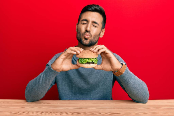 Pohledný hispánec jí chutný klasický burger, dívá se do kamery a posílá polibek, který je krásný a sexy. výraz lásky.  - Fotografie, Obrázek