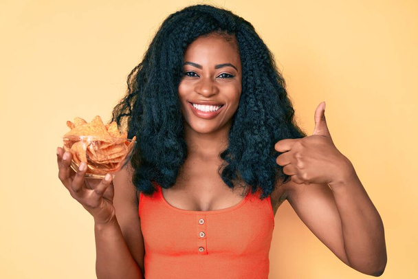Beautiful african woman holding nachos potato chips smiling happy and positive, thumb up doing excellent and approval sign  - Foto, Bild