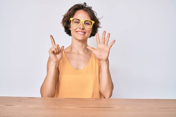 Young hispanic woman wearing casual clothes and glasses sitting on the table showing and pointing up with fingers number seven while smiling confident and happy.  - Foto, Imagem
