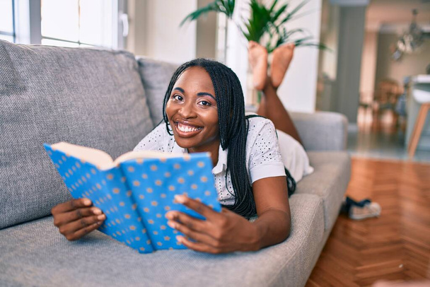 Junge afrikanisch-amerikanische Frau lächelt glücklich auf dem Sofa liegend und liest Buch zu Hause - Foto, Bild