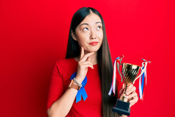 Giovane donna cinese che indossa il primo posto distintivo che tiene il trofeo faccia seria pensando alla domanda con mano sul mento, riflessivo sull'idea confusa  - Foto, immagini