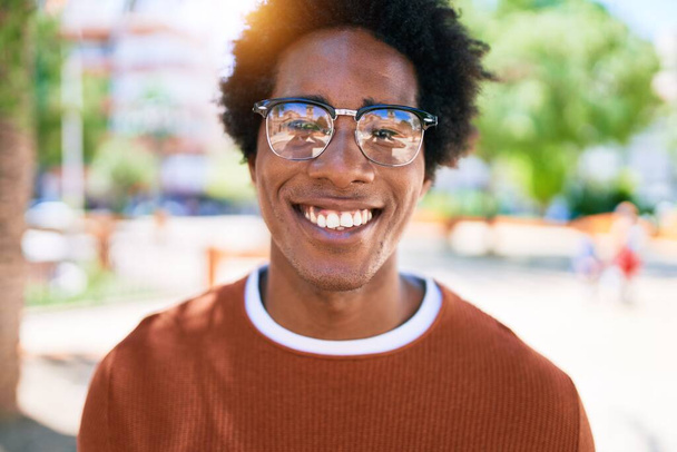Młody, przystojny, afrykański mężczyzna w luźnych ubraniach i okularach uśmiechnięty szczęśliwy. Stojąc z uśmiechem na twarzy patrząc w kamerę idącą ulicą miasta. - Zdjęcie, obraz