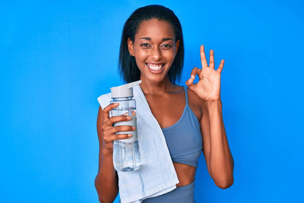 Nuori afrikkalainen amerikkalainen nainen yllään urheiluvaatteet vesipullo tekee ok merkki sormilla, hymyillen ystävällinen ele erinomainen symboli  - Valokuva, kuva