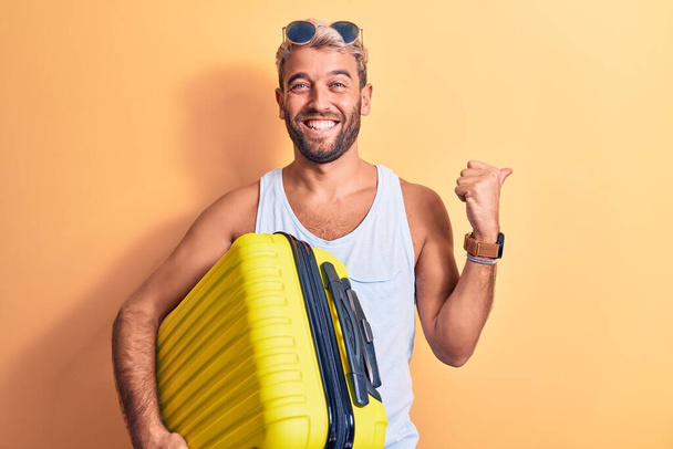 Young handsome blond tourist man on vacation wearing sunglasses holding aiplane cabin bag pointing thumb up to the side smiling happy with open mouth - Photo, image