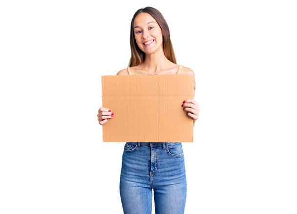 Красива брюнетка молода жінка тримає картонний банер з порожнім простором виглядає позитивно і щасливо стоячи і посміхаючись з впевненою посмішкою, показуючи зуби
  - Фото, зображення