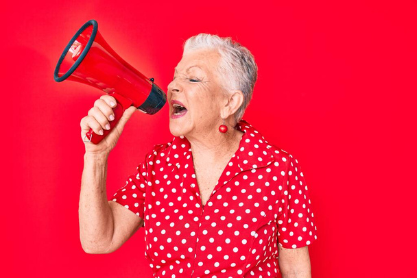 Senior hermosa mujer de pelo gris gritando usando megáfono sobre fondo rojo aislado - Foto, Imagen