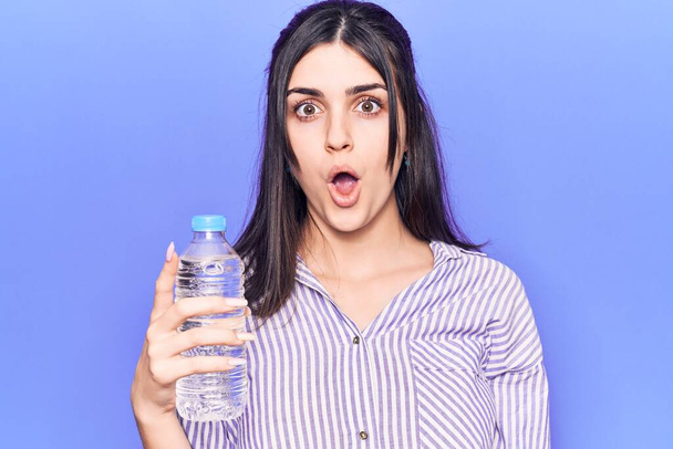 Young beautiful girl holding bottle of water scared and amazed with open mouth for surprise, disbelief face  - Photo, Image