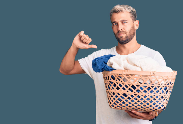 Jonge knappe blonde man houdt wasmand met kleren met boos gezicht, negatief teken toont een hekel aan met duimen naar beneden, afwijzing concept  - Foto, afbeelding