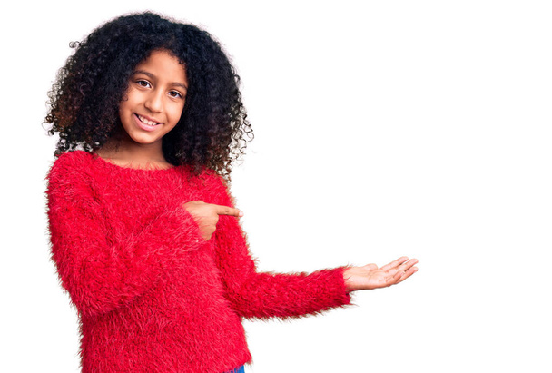 Afrikaans amerikaans kind met krullend haar dragen casual winter trui verbaasd en glimlachen naar de camera terwijl presenteren met de hand en wijzen met de vinger.  - Foto, afbeelding