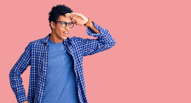 Giovane uomo afroamericano indossa abiti casual e occhiali molto felice e sorridente guardando lontano con mano sopra la testa. concetto di ricerca.  - Foto, immagini