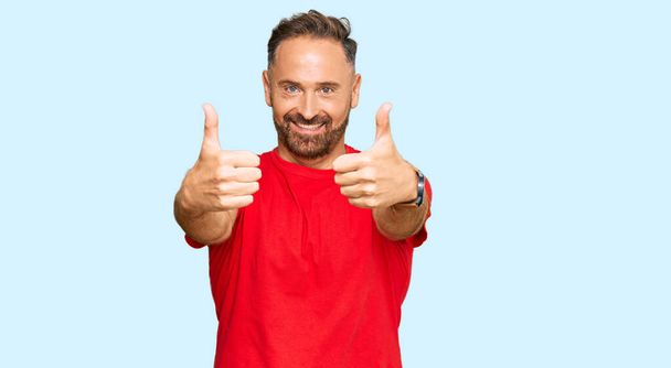 Hombre guapo de mediana edad que usa una camiseta roja casual que aprueba hacer un gesto positivo con la mano, pulgares arriba sonriendo y feliz por el éxito. gesto ganador.  - Foto, Imagen