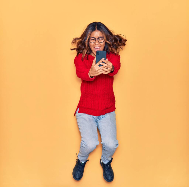 Jovem bela mulher latina usando óculos sorrindo feliz. Saltar com sorriso no rosto fazer selfie por smartphone sobre fundo amarelo isolado - Foto, Imagem
