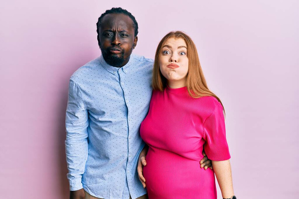 Young interracial couple expecting a baby, touching pregnant belly puffing cheeks with funny face. mouth inflated with air, crazy expression.  - Fotó, kép