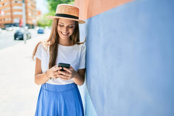Tatildeki genç sarışın kadın mutlu bir şekilde gülümsüyor. Akıllı telefon kullanıyor ve şehrin sokaklarında duvara yaslanıyor. - Fotoğraf, Görsel