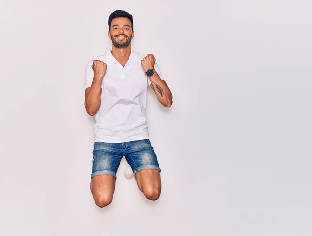 Young handsome hispanic man wearing casual clothes smiling happy. Jumping with smile on face celebrating with fists up over isolated white background - Foto, Imagem