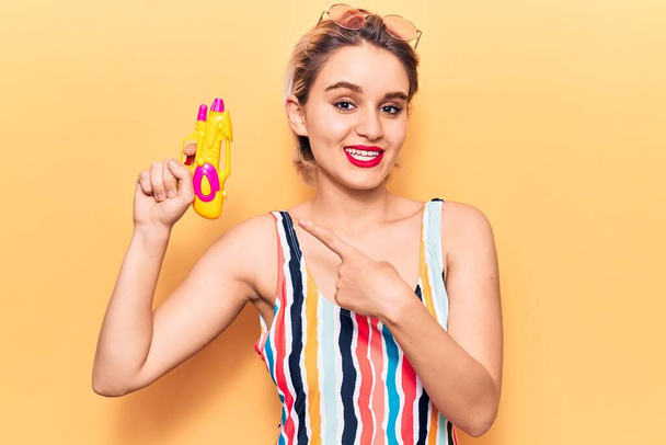 Young beautiful blonde woman wearing swimwear holding water gun smiling happy pointing with hand and finger  - Photo, Image