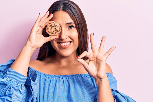 Young beautiful brunette woman holding cookie doing ok sign with fingers, smiling friendly gesturing excellent symbol  - Foto, Imagen