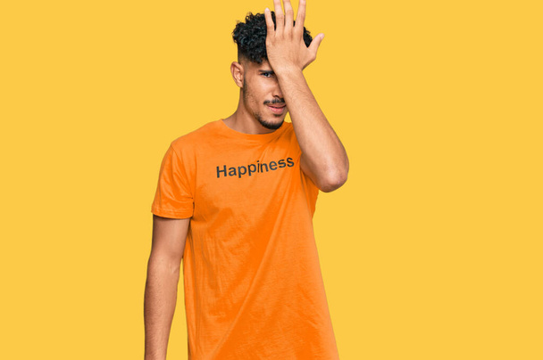 Νεαρός Άραβας φοράει μπλουζάκι με μήνυμα ευτυχίας λέξη έκπληκτος με το χέρι στο κεφάλι για λάθος, θυμηθείτε λάθος. ξεχασμένη, κακή έννοια μνήμης.  - Φωτογραφία, εικόνα