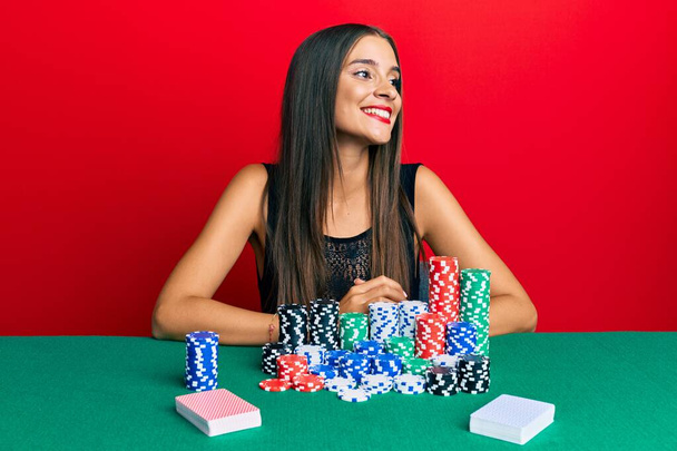 Giovane donna ispanica seduta sul tavolo a giocare a poker guardando da un'altra parte con sorriso sul viso, espressione naturale. ridendo fiducioso.  - Foto, immagini