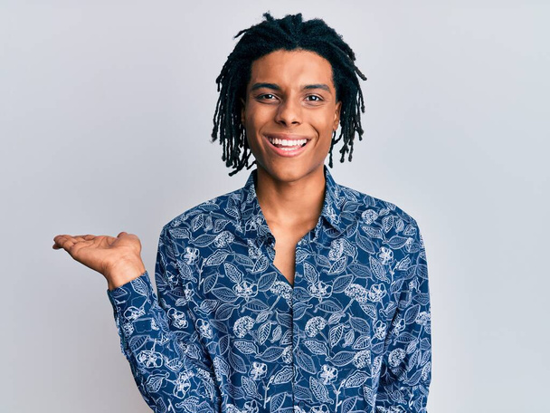 Young african american man wearing 80s style shirt smiling cheerful presenting and pointing with palm of hand looking at the camera.  - Photo, Image