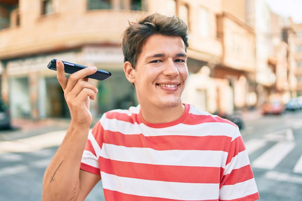 Młody biały mężczyzna uśmiecha się szczęśliwy słuchając wiadomości audio za pomocą smartfona w mieście. - Zdjęcie, obraz