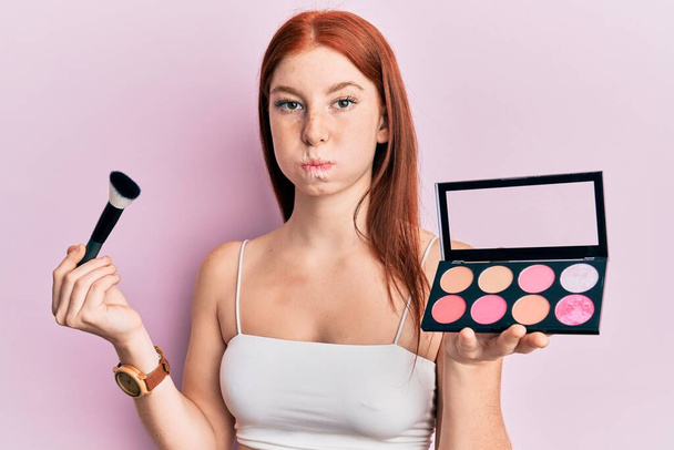 Young red head girl holding makeup brush and blush palette puffing cheeks with funny face. mouth inflated with air, catching air.  - Photo, Image