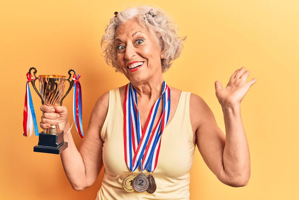 Senior grey-haired woman holding champion trophy wearing medals celebrating achievement with happy smile and winner expression with raised hand  - Zdjęcie, obraz