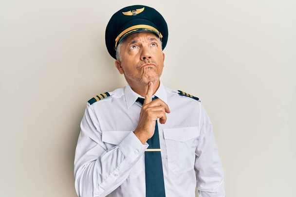 Schöner reifer Mann mittleren Alters in Flugzeugpilotenuniform, der konzentriert über Zweifel nachdenkt, mit dem Finger am Kinn nach oben schaut und sich wundert  - Foto, Bild