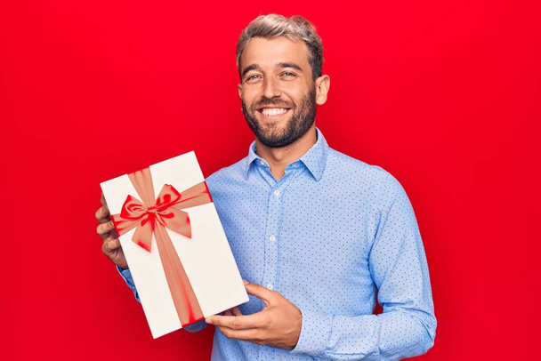 Joven hombre rubio guapo con barba sosteniendo regalo de cumpleaños sobre fondo rojo aislado mirando positiva y feliz de pie y sonriendo con una sonrisa confiada mostrando los dientes - Foto, imagen