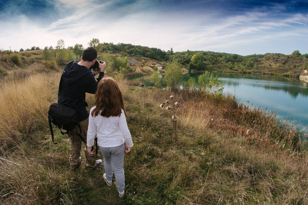 Laaja taustakuva valokuvaajasta ja pikkutytöstä kuvaamassa kohtausta kauniilla järvellä pilvisenä ja syyspäivänä. Vanhemmuuden käsite - Valokuva, kuva