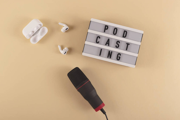 Podcast-Schriftzug neben Mikrofon und drahtlosen Kopfhörern am Arbeitsplatz auf beigem Hintergrund. - Foto, Bild