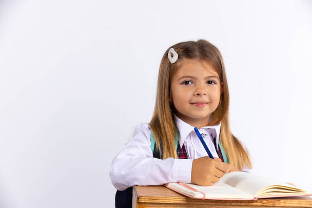 Handsome little girl in school uniform sitting at desk isolated, white background with free space . Happiest schoolgirl doing homework. Education concept - Valokuva, kuva