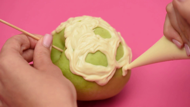 partial view of woman drawing scary face with cream on ripe apple on pink - Footage, Video