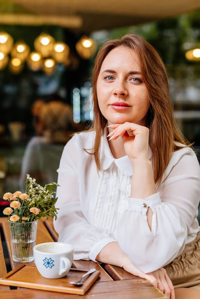 Young beautiful woman holding a cup of coffee in cafe. Outdoor portrait of female blogger posing while traveling holiday vacation. Happy freelancer thinking about new ideas. Looking at camera - Fotó, kép