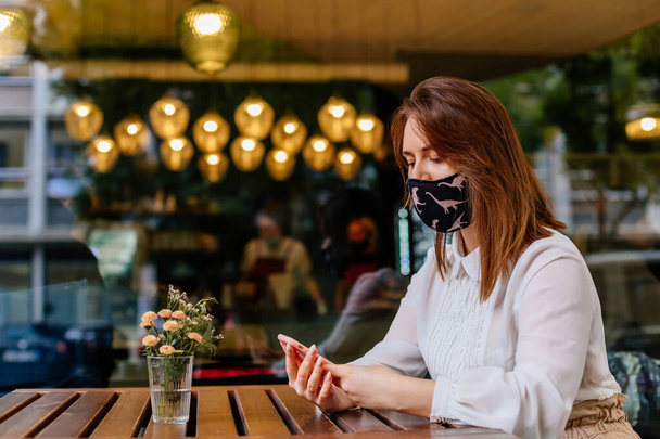 Kafkasyalı, koruyucu maske takan bir iş kadını bir kafede dizüstü bilgisayarla çalışıyor. COVID 19 virüs koruması. Uzaktan çalışma ve internetten öğrenme kavramı. Bilgisayara bakıyorum. - Fotoğraf, Görsel