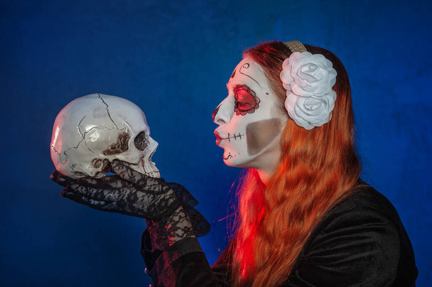 Beautiful woman with scary Halloween make up dead day calavera style kissing human skull on blue backgroun - Foto, imagen