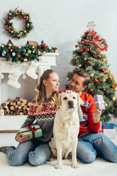 χαρούμενο ζευγάρι κρατώντας δώρα και κάθεται κοντά σε σκύλο και χριστουγεννιάτικο δέντρο - Φωτογραφία, εικόνα