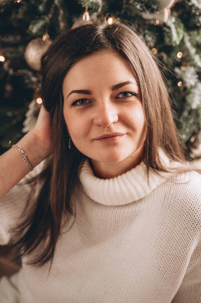 Portrait of a brunette girl in a white sweater near the Christmas tree - Fotoğraf, Görsel