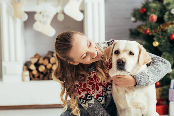 happy woman in sweater looking at labrador near christmas tree and fireplace - Foto, Imagen