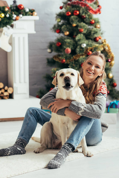 šťastná žena ve svetru sedí na podlaze s labrador v blízkosti vánočního stromku  - Fotografie, Obrázek