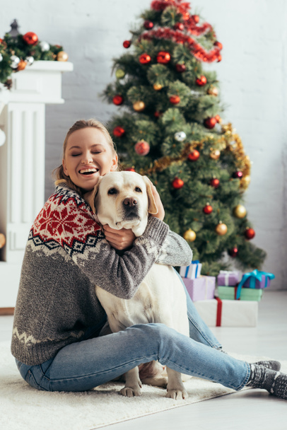 femme heureuse avec les yeux fermés assis sur le sol et embrasser labrador près de l'arbre de Noël  - Photo, image