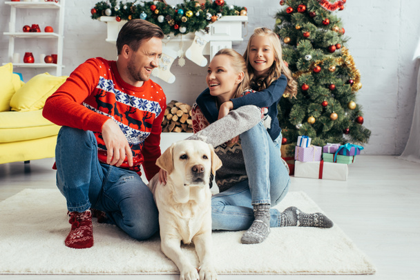 familia feliz en suéteres abrazando labrador cerca del árbol de navidad decorado  - Foto, imagen