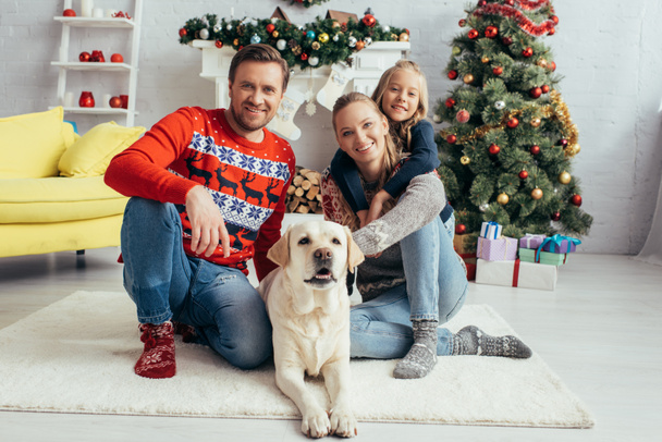 happy parents in sweaters cuddling labrador near daughter and decorated christmas tree - Photo, Image