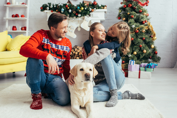 子供の近くにラブラドールを抱きかかえてクリスマスツリーを飾るセーターの幸せな両親 - 写真・画像